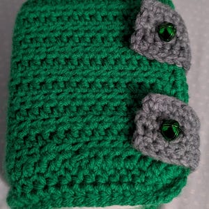 May include: A handmade, green crocheted item with two gray, bell-adorned accents. The item is made of a thick, textured yarn, and the bells are a shiny green color. The overall design is simple and cozy.
