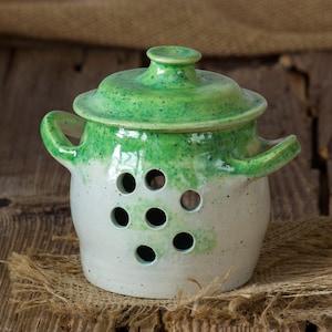 Könnte beinhalten: Ein weißer Keramik-Knoblauchtopf mit grünem Deckel und Belüftungslöchern. Der Topf hat eine gesprenkelte grüne Glasur oben und eine weiße Glasur unten.