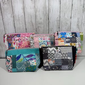May include: A collection of five quilted zipper pouches in various sizes and patterns. The pouches feature patchwork designs with floral, geometric, and heart motifs. Colors include pink, blue, green, and gray. One pouch has the text "You are my sun".