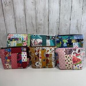May include: A collection of six handmade zippered pouches. Each pouch features a patchwork design with various fabric patterns and colors, including floral, geometric, and paisley prints. The pouches have a rectangular shape and a zipper closure.