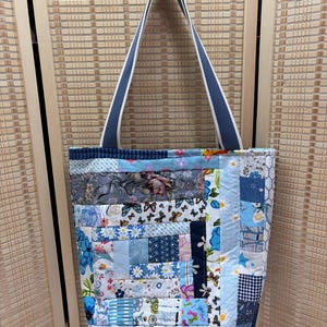 May include: A rectangular tote bag constructed from a patchwork of various blue, white, and grey fabric squares featuring floral, butterfly, and geometric patterns. The bag includes two long, solid blue shoulder straps attached to the top edge.