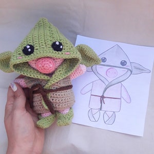 Puede incluir: Un juguete de peluche tejido a crochet con forma de cerdo que lleva una bata con capucha verde. El juguete está siendo sostenido en la mano de una persona. Un dibujo de un cerdo similar con una bata se muestra en el fondo.