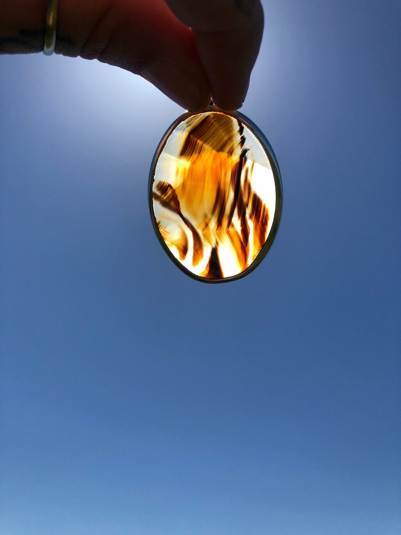 May include: A brown and white agate pendant with a silver setting. The pendant is oval shaped and has a swirling pattern.