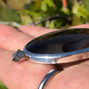 May include: A silver pendant featuring a dark brown agate stone. The pendant is oval shaped and has a smooth, polished finish. The stone displays a natural, organic pattern. The pendant is marked with "925" on the side.