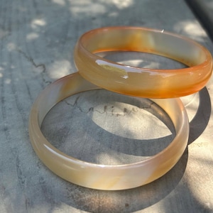 May include: Two smooth, polished bangle bracelets made of translucent stone. The top bangle features a vibrant orange hue, while the bottom bangle displays a lighter, creamy off-white color with subtle, soft orange swirls throughout the material.
