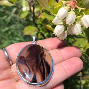 May include: A silver pendant featuring a brown and white agate stone. The stone displays a distinctive swirling pattern of lines. The pendant is oval shaped and has a smooth, polished finish.