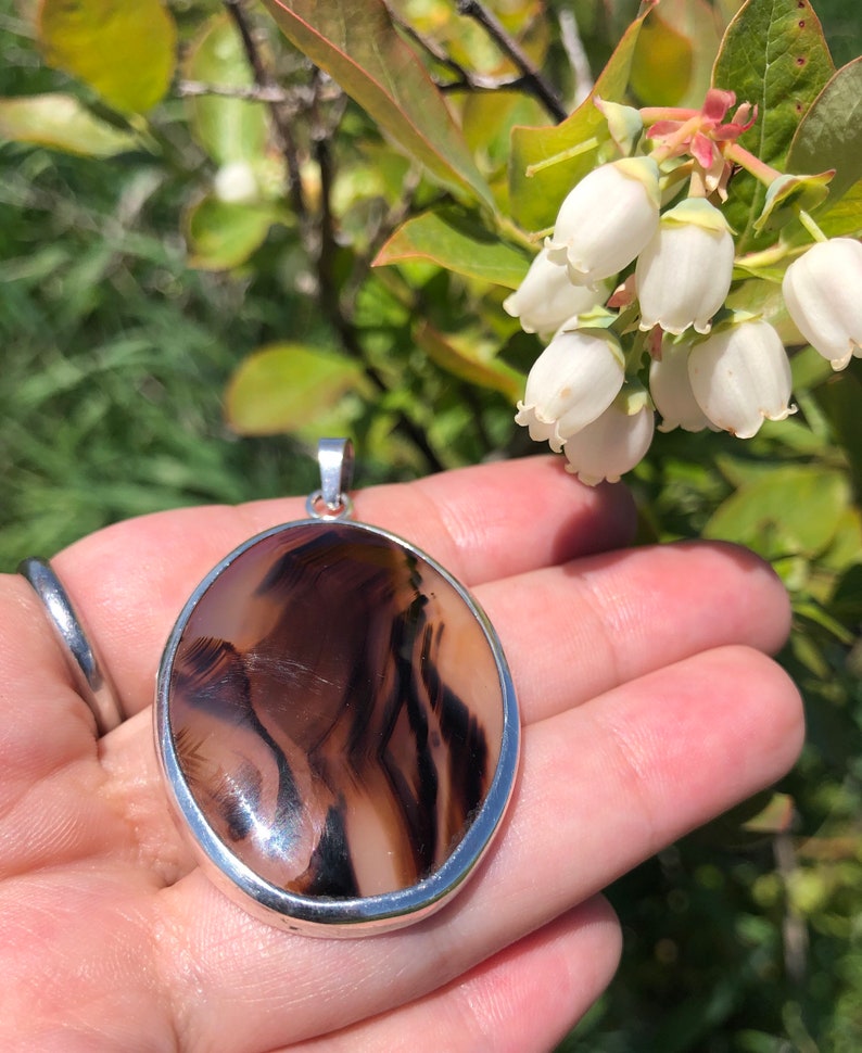 May include: A silver pendant featuring a brown and white agate stone. The stone displays a distinctive swirling pattern of lines. The pendant is oval shaped and has a smooth, polished finish.