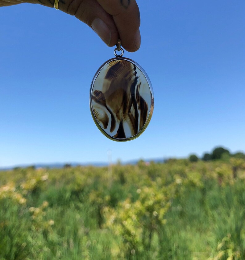 May include: A silver oval pendant featuring a brown and white agate stone. The stone has a swirling pattern.
