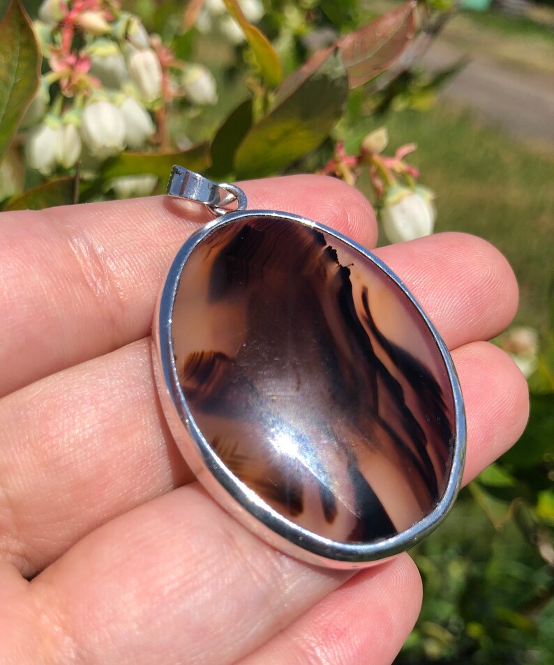 May include: A silver pendant featuring a brown and white agate stone. The stone displays a distinctive pattern of swirls and lines.