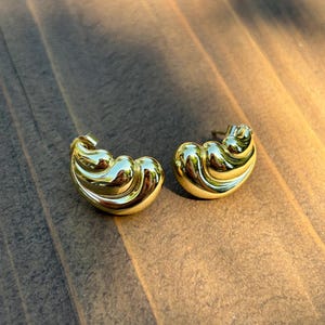 May include: A pair of gold-toned earrings featuring a curved, multi-layered shell-like design with a polished, reflective surface. Each earring has a smooth, rounded silhouette with distinct ridges that create a sense of depth and volume.