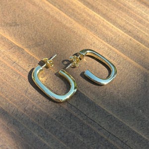 May include: A pair of gold-toned hoop earrings with a rectangular, rounded-corner shape. Each earring features a post-back closure and a smooth, polished metallic finish. The interior of one earring is clearly stamped with the text AV 925.
