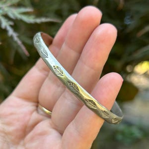 May include: A silver-toned metal bangle bracelet featuring a repeating engraved pattern of small, stylized leaves along the outer surface. The bracelet has a smooth, polished finish and a rounded profile, showcasing a consistent design throughout.