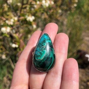 May include: A silver ring with a large, teardrop-shaped green malachite stone. The stone has a swirling pattern of dark and light green.
