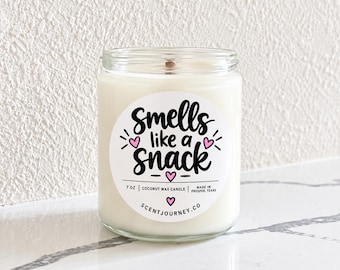 May include: A clear glass jar candle filled with white coconut wax and a wooden wick. The circular label features the black text "smells like a snack" with small pink hearts, "7 oz | coconut wax candle", and "scentjourney.co" printed on it.