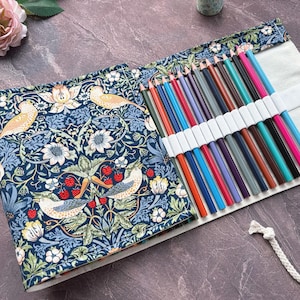 Peut inclure: Trousse à crayons ouverte, enroulée, avec un assortiment de crayons de couleur. La trousse présente un motif floral bleu avec des oiseaux et des fraises. Un cordon blanc est attaché pour la fermeture.