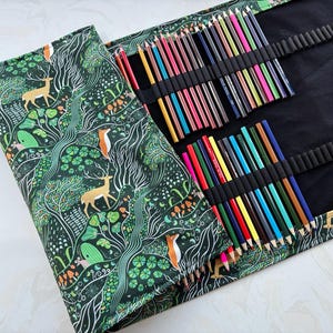 Peut inclure: Une trousse à crayons en tissu avec un imprimé forêt vert foncé représentant des cerfs, des renards et des champignons. L'intérieur noir comporte des boucles élastiques contenant de nombreux crayons de couleur. Des crayons sont posés à côté.