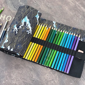 Puede incluir: Un estuche negro para lápices con un estampado de olas en azul y dorado contiene una variedad de lápices de colores. Los lápices están ordenados por color, de amarillo a morado. Se ven algunos lápices sueltos.