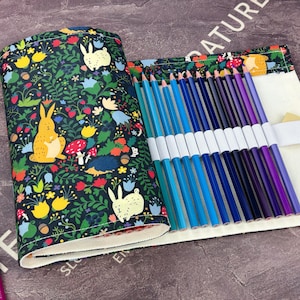 以下が含まれることがあります： ウサギ、キノコ、花が描かれた、気まぐれな森の模様のロールペンケース。ケースは開いており、青と紫の色鉛筆のセットが表示されています。いくつかの緩い鉛筆が見えます。