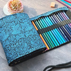 Könnte beinhalten: Eine blaue Stiftrolle mit floralem Muster, geöffnet, um eine Sammlung von Farbstiften in verschiedenen Farbtönen zu zeigen. Die Rolle ist aus Stoff und hat einen schwarzen Riemen. Eine rosa Blume und andere Kunstutensilien sind im Hintergrund.