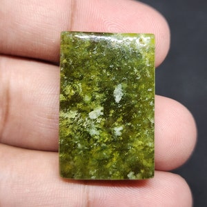 May include: A green and white gemstone cabochon with a rectangular shape. The stone has a mottled appearance with green and white inclusions.