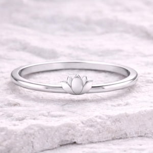Peut inclure: Une bague en argent ornée d'une fleur de lotus délicate. La bague a un anneau fin et une pièce maîtresse florale détaillée. La bague est présentée sur une surface blanche texturée, soulignant son élégance. Un bijou.