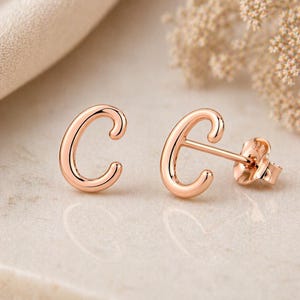 May include: Rose gold letter "C" stud earrings. The earrings are in the shape of the letter "C" with a curved design. The earrings are on a light-colored surface with a soft, blurred background.