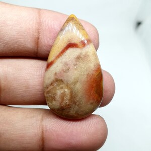 May include: A teardrop-shaped gemstone with a polished surface. The stone features a blend of colors, including beige, orange, and red, with a layered pattern. The gemstone is held in a hand, showcasing its size and shape.