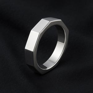 Könnte beinhalten: Ein silberner Ring mit achteckigem Design. Der Ring hat eine polierte, reflektierende Oberfläche und ist vor einem dunklen Hintergrund platziert. Die Form des Rings ist kantig und modern. Das Design des Rings ist geometrisch.