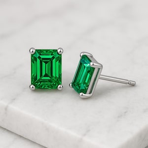 Peut inclure: Une paire de boucles d'oreilles à clous en forme d'émeraude verte avec un sertissage argenté. Les boucles d'oreilles sont présentées sur une surface blanche, mettant en valeur leur couleur vive et leur design géométrique.