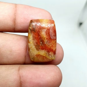 Könnte beinhalten: Ein rechteckiger Edelstein mit polierter Oberfläche. Der Stein weist eine Mischung aus Farben auf, darunter Rot, Orange und Gelb, mit einem marmorierten Muster. Der Edelstein wird zwischen den Fingern gehalten, um seine Größe und Form zu zeigen.