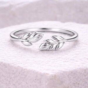 May include: A silver ring with an open design, featuring two detailed leaf motifs. The ring is set against a white background, highlighting the intricate design and metallic sheen. A delicate piece of jewellery.