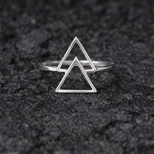 May include: A silver ring with a double triangle design. The ring sits on a dark, textured surface. The triangles are open, and the band is slender. The ring's design is geometric.