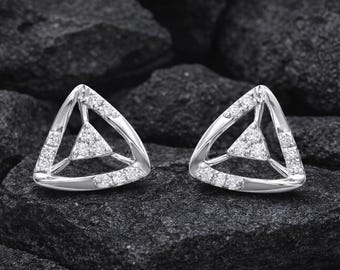 May include: A pair of silver-toned triangle stud earrings. Each earring features a larger outer triangle with a smaller inner triangle, both encrusted with small, sparkling gemstones. The earrings are displayed on a dark, textured surface.
