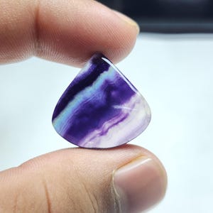 Puede incluir: Un cabujón de fluorita púrpura y blanco con forma de lágrima. La piedra tiene un patrón de remolino de púrpura y blanco.