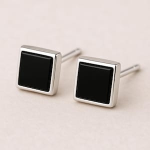 May include: A pair of square stud earrings with a black center and a silver-colored border. The earrings have a post backing. The earrings are displayed on a light beige surface.