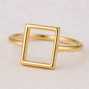 May include: A gold ring with a square geometric design. The ring has a slender band and a hollow square at the centre. The ring is made of a shiny, polished metal. The ring is displayed on a white surface.
