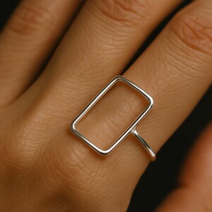 May include: A silver ring with a rectangular open frame design. The ring is worn on a finger, highlighting its minimalist and modern style. The ring's clean lines and geometric shape create a contemporary appearance.