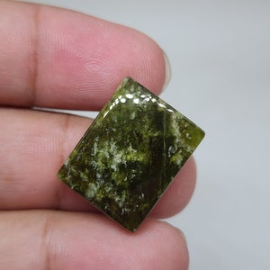 May include: A rectangular gemstone with a mix of green and white hues. The stone has a polished surface and a natural, organic pattern. The gemstone is held between fingers, showcasing its size and shape.