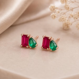 May include: A pair of stud earrings in rose gold, each featuring a rectangular emerald-cut red gemstone paired with a teardrop-shaped green gemstone. The stones are set side-by-side in a metal frame with a post backing for secure attachment.