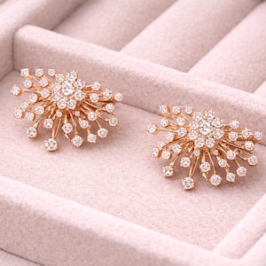 May include: Two gold-toned, starburst-shaped earrings with numerous small, sparkling clear gemstones. The earrings are displayed in a light pink jewelry box with a soft, velvety texture. The design is symmetrical and elegant.