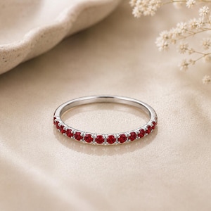 May include: A thin silver-toned band ring featuring a row of small, round, vibrant red gemstones set along the front half of the metal band. The ring has a polished metallic finish and a consistent, delicate design with evenly spaced stones.