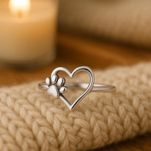 Peut inclure: Bague en argent avec un motif en forme de cœur et une empreinte de patte. La bague est posée sur un fond doux et texturé, avec une bougie floue en arrière-plan. La bague est un symbole d'amour pour les animaux.