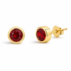 May include: A pair of gold stud earrings with round, vibrant red gemstones. The earrings have a simple, elegant design, with the red stones set in a gold bezel. The post and backing are also gold. The earrings are displayed on a white background.