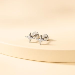 May include: A pair of silver-colored star-shaped stud earrings. Each earring features a four-pointed star design with a post and backing. The earrings are displayed on a light beige surface.
