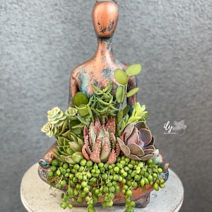 May include: A copper-colored, distressed, ceramic figurine of a person in a seated yoga pose, with a variety of succulents planted in the figure's lap. The succulents include string of pearls, echeveria, and other varieties.