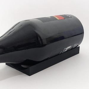 Puede incluir: Una botella de vino negra apoyada sobre un soporte rectangular negro. La botella es oscura con una etiqueta con detalles rojos y dorados. El soporte tiene un recorte circular para sostener la botella.