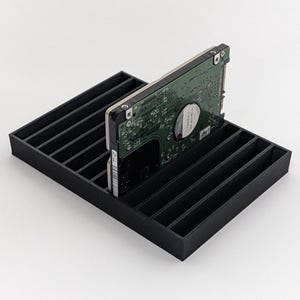 Puede incluir: Un organizador de disco duro negro impreso en 3D con múltiples ranuras, que sostiene un disco duro de placa de circuito verde. El organizador es rectangular y está diseñado para guardar varios discos duros, manteniéndolos en posición vertical y organizados.