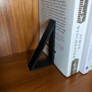 Op de afbeelding: Een zwarte driehoekige boekenlegger met een gestructureerd oppervlak. De boekenlegger houdt twee boeken vast, een met de titel "Simon Winchester" en de andere met de titel "Tilly".