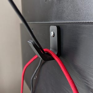 May include: A black plastic hook mounted on a black surface with a red and black cord wrapped around it.