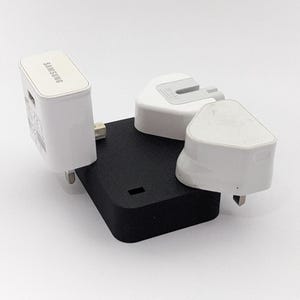 Può includere: Una stazione di ricarica nera con tre adattatori di alimentazione bianchi. Un adattatore è etichettato "SAMSUNG". La stazione ha una forma rettangolare con bordi arrotondati e un piccolo ritaglio rettangolare.